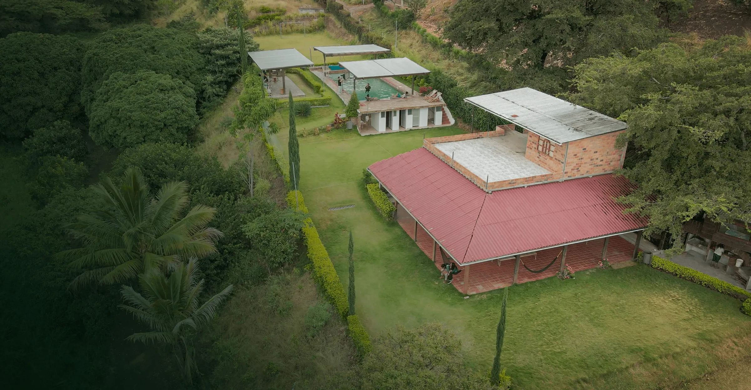 Vista aérea de la granja: casa de techo rojo con corredor y hamacas, piscina techada, quioscos y cancha entre árboles